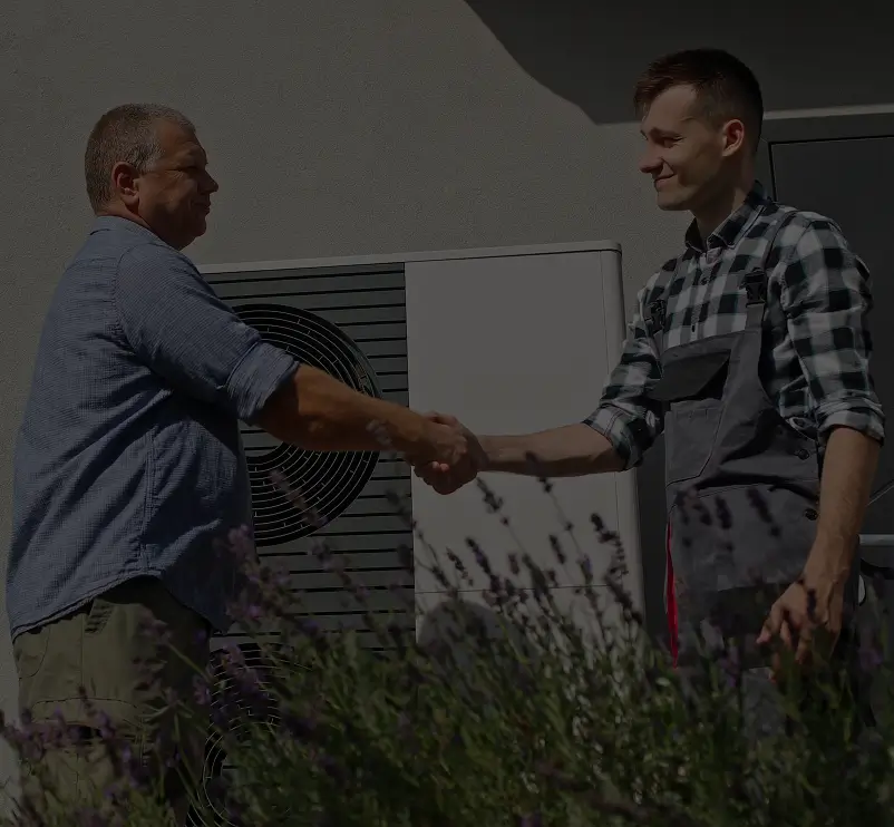 Two men shaking hands in front of an outdoor unit, one in a plaid shirt and the other in overalls, with plants in the foreground.