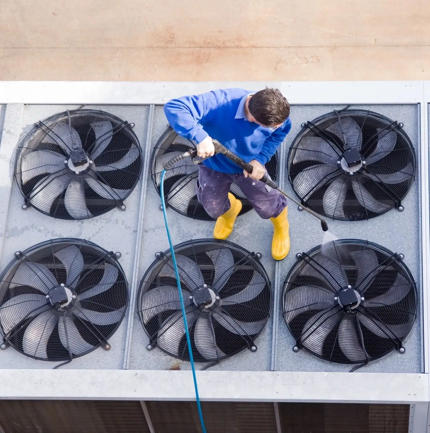 Person in blue shirt and yellow boots pressure washing large industrial fans on a rooftop.